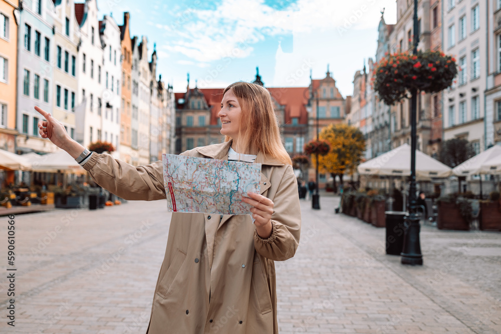 Happy young woman tourist with trendy look with map visiting city and looking around, searching direction on location map during vacation in Gdansk old town.
