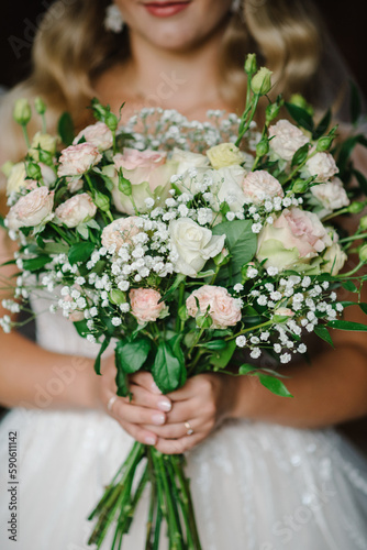 Young bride in elegant dress is standing and holding hand bouquet of pink flowers and greens at nature.