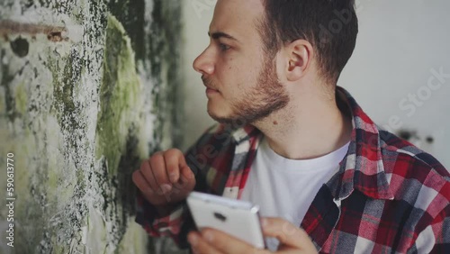 a man in a shirt with a phone in his hands examines a wall with mold ,searching the Internet for a disinfection service close up