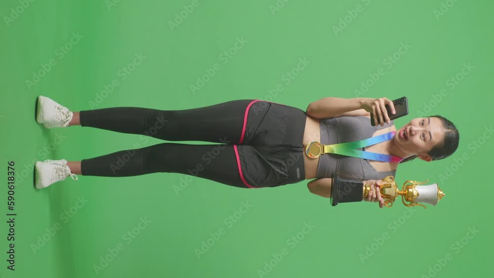 Full Body Of Asian Woman With A Gold Medal And Trophy Looking At Smartphone Then Screaming Goal Celebrating On Green Screen Background In The Studio
