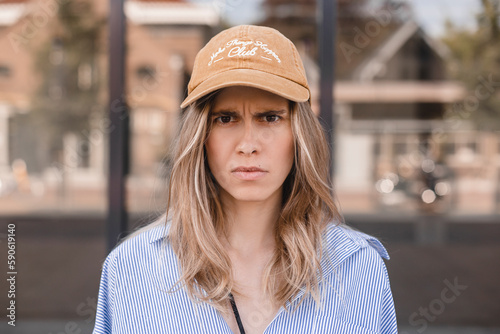 Drama blonde curly woman look angry at camera walking on the street. Girl wear beige cap and stripped shirt. Portrait of a lovely angry young girl wearing casual clothing standing.