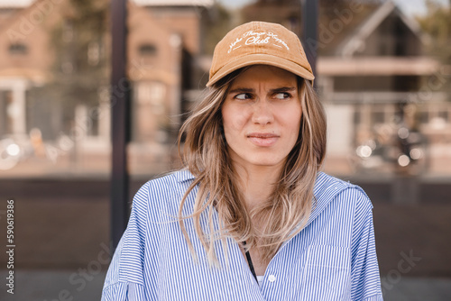 Evil blonde curly woman look angry at camera walking on the street. Girl wear beige cap and stripped shirt. Portrait of a disgusted angry young girl wearing casual clothing standing near office.