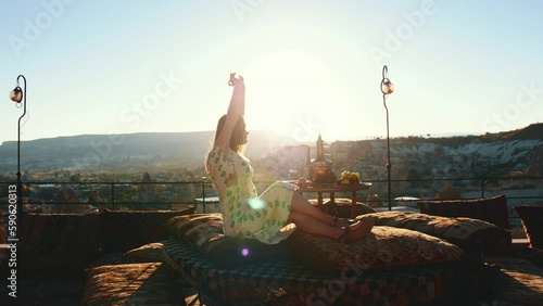 Happy young female stretching and enjoying breakfast during sunrise on authentic terarace in Cappadocia. Panoramic view