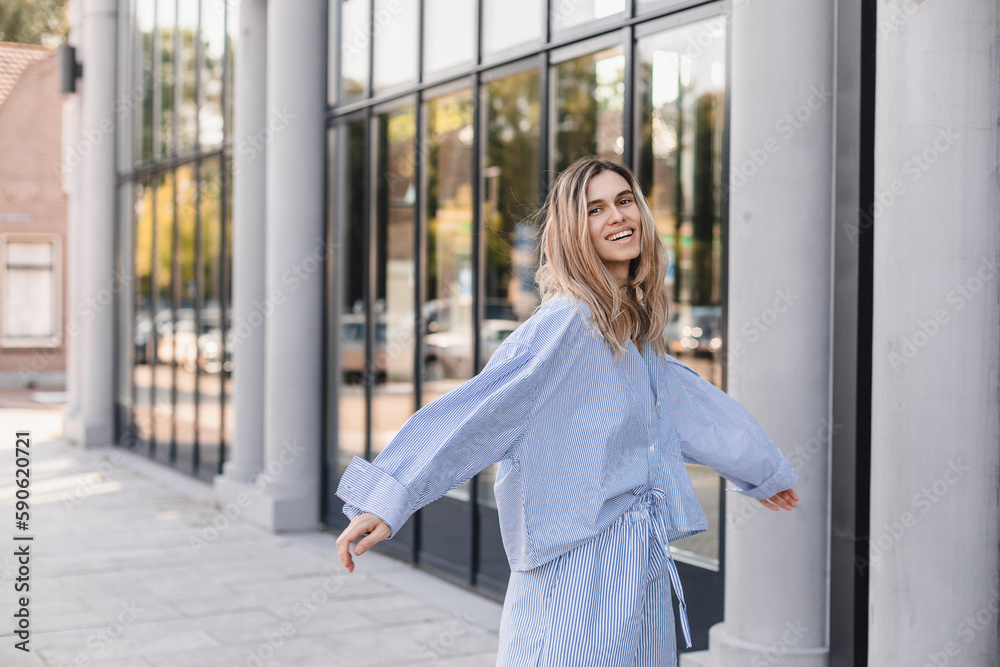 Attractive beauty woman walking on the street while wearing stripped shirt. Blonde girl is turn around near office building. Mirror windows. Female fix her hair while walk in the city.