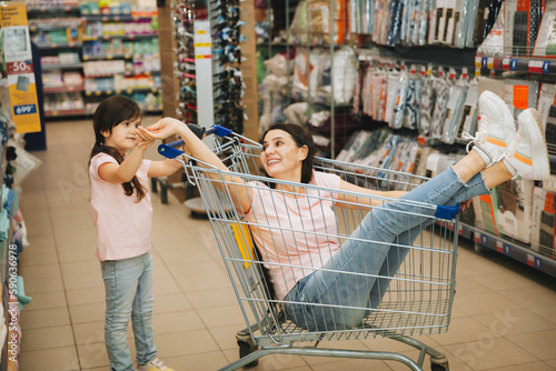 Εκτύπωση καμβά In the supermarket: a daughter pushes a cart with a woman sitting in it, a happy family rushes cheerfully on the cart in the store