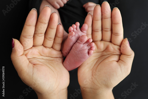The palms of the parents. A mother hold the feet of a newborn child in a black blanket on a Black background. The feet of a newborn in the hands of parents. Macro Photo of foot, heels and toes