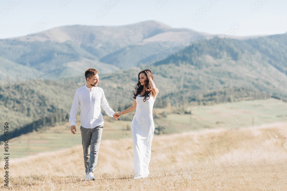 A stylish model couple in the mountains in the summer. A young boy and a girl in a white silk dress are walking on the slope against the background of the forest and mountain peaks. Portrait.
