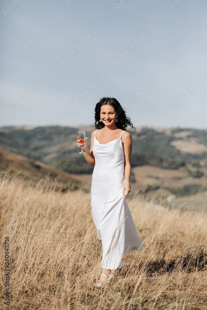 A beautiful girl with beautiful black hair in a white dress walks smiling at the camera with a glass of champagne, her long dress with the other hand. Portrait of a girl. A beautiful girl with beautiful black hair in a white dress walks smiling at the camera with a glass of champagne, her long dress with the other hand. Portrait of a girl.