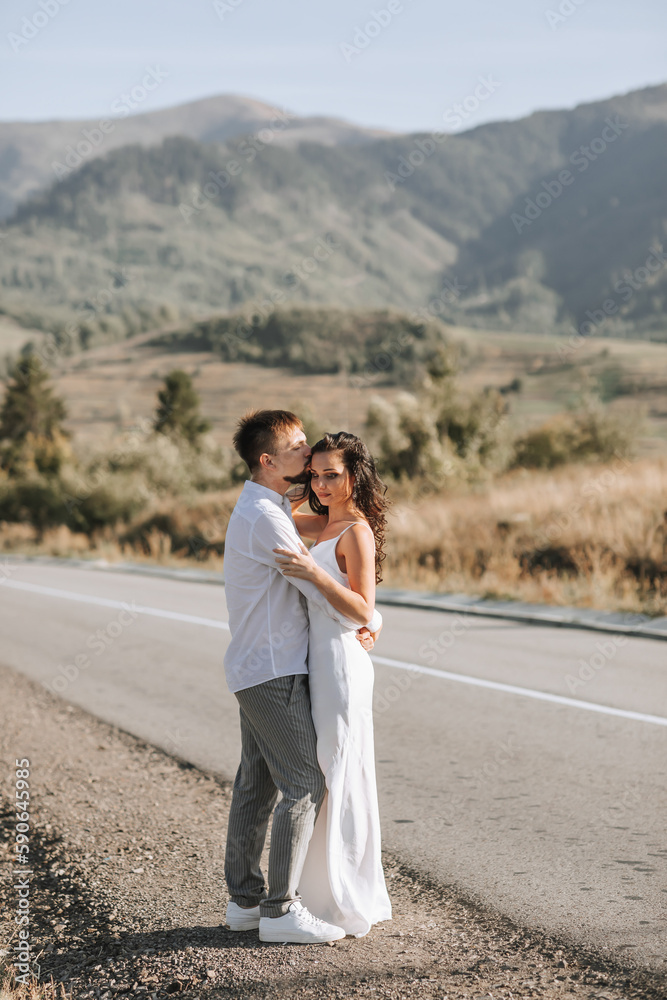 A stylish groom in a white shirt and a cute brunette bride in a white dress are hugging on the side of the road against the background of the forest and mountains. Wedding portrait of newlyweds. A stylish groom in a white shirt and a cute brunette bride in a white dress are hugging on the side of the road against the background of the forest and mountains. Wedding portrait of newlyweds.