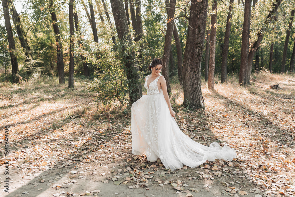 The bride in a white dress with a long train is walking in the forest, looking down. The wind blows the girl's hair. The shadows Summer wedding. The bride in a white dress with a long train is walking in the forest, looking down. The wind blows the girl's hair. The shadows Summer wedding.