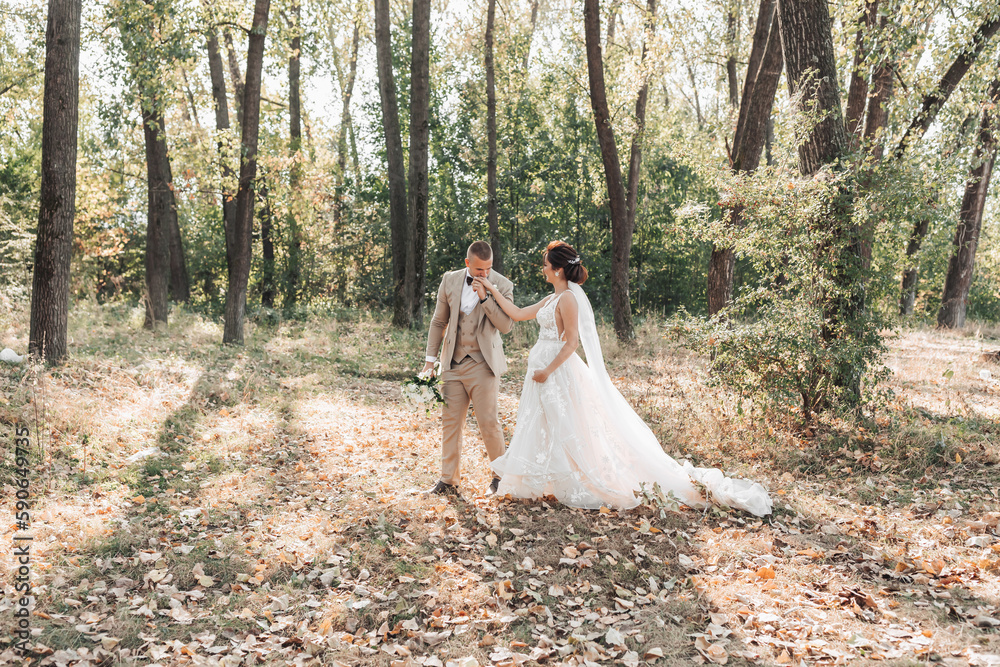 Wedding photo. The bride and groom are walking in the forest. The groom holds a bouquet and kisses the hand of his beloved. Long wedding dress. Couple in love. Summer light. The shadows