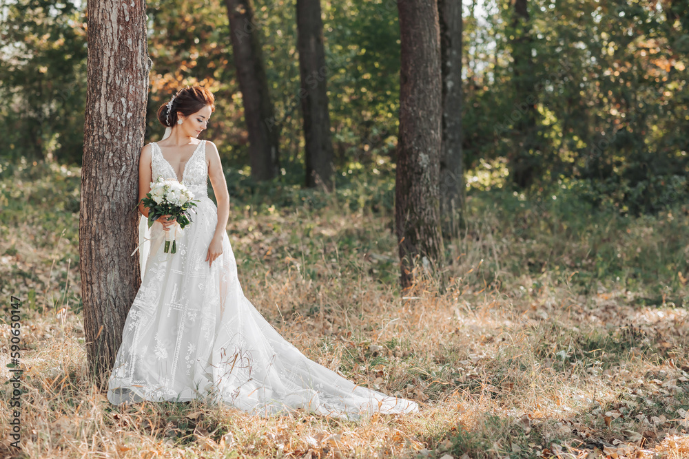 Wedding photo in nature. The bride is standing near a tree in a beautiful dress, holding her bouquet of white roses, looking to the side. Beautiful makeup of the bride. Portrait. Wedding photo in nature. The bride is standing near a tree in a beautiful dress, holding her bouquet of white roses, looking to the side. Beautiful makeup of the bride. Portrait.