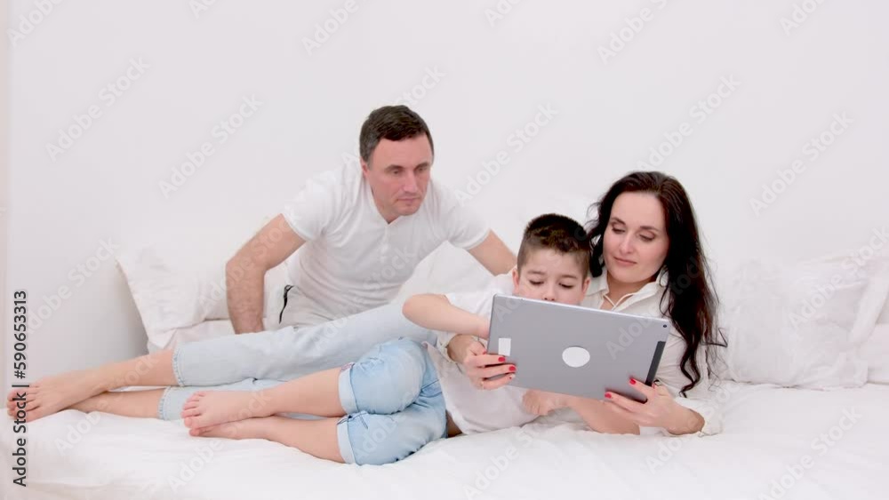 family in white bed with tablet examining game father showing how to press buttons correctly mother and son smiling laughing rejoicing child learning new game developing lessons cheerful family love family in white bed with tablet examining game father showing how to press buttons correctly mother and son smiling laughing rejoicing child learning new game developing lessons cheerful family love