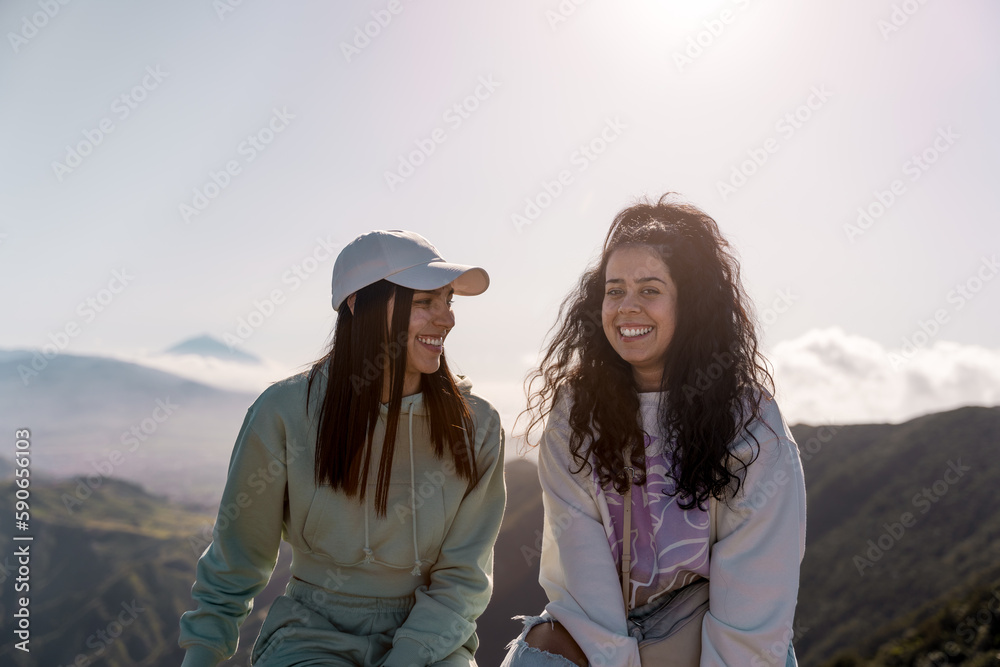 Mujer latina mirando a mujer europea con una gran sonrisa mirando a cámara con montañas desenfocado de fondo, tonos de color cálidos 