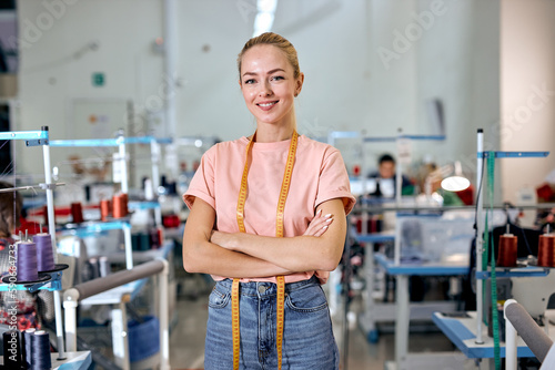 female designer of clothes standing among sewing machines in textile factory, attractive tailor after work stand posing, looking at camera smiling with arms crossed, measuring tape for sewing on neck