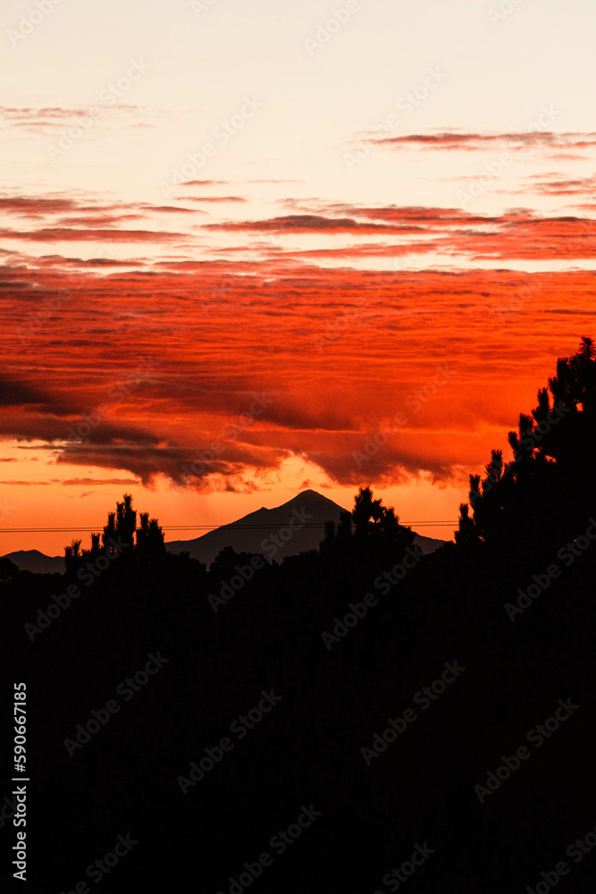 Obraz premium Sunrise with the sun's rays leaving the horizon, reflecting in the clouds and in the background a volcano.