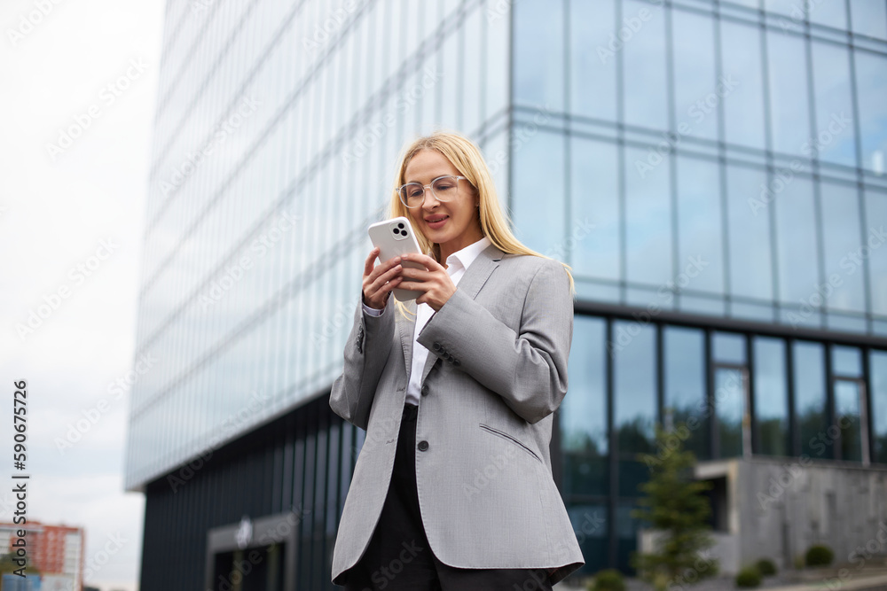 Beautiful smiling businesswoman wearing stylish eyeglasses using mobile phone chatting, communication online standing on the street, selective focus. Technology concept Beautiful smiling businesswoman wearing stylish eyeglasses using mobile phone chatting, communication online standing on the street, selective focus. Technology concept