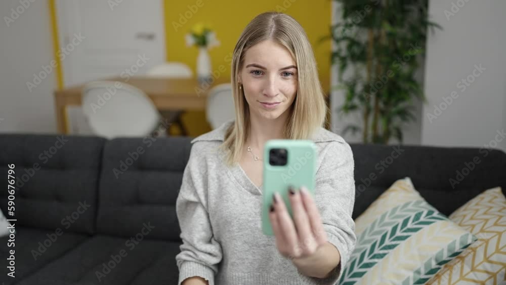 Young blonde woman doing video call with phone sitting on the sofa at home