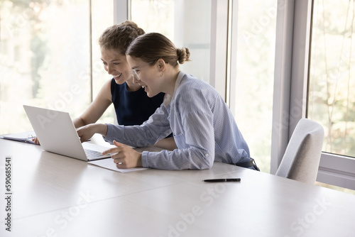 Two cheerful young female employees sharing workplace, using online application on laptop, watching presentation on Internet together, smiling, laughing