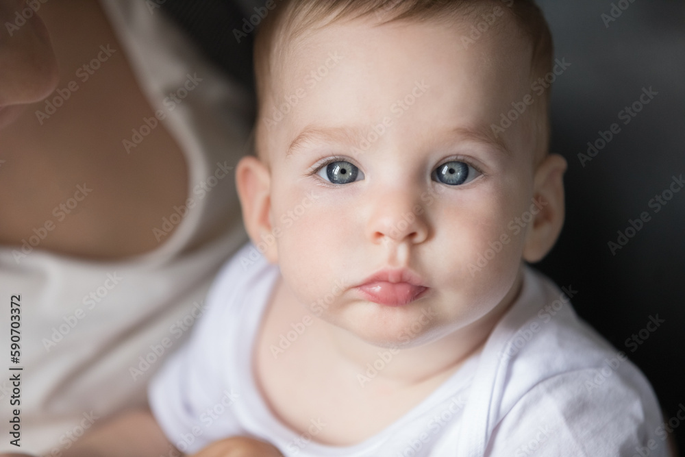 Face of pretty adorable blue eyed baby held by mom looking at camera. New mother holding little kid with tenderness, love, care, enjoying childcare. Beautiful infant indoor closeup portrait