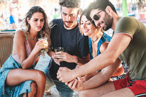 Caucasian Friends Enjoying Beach Bar- Four friends, two men and two women, with various summer outfits, laughing and looking at a bearded man's smartphone while enjoying mojitos. © Lomb
