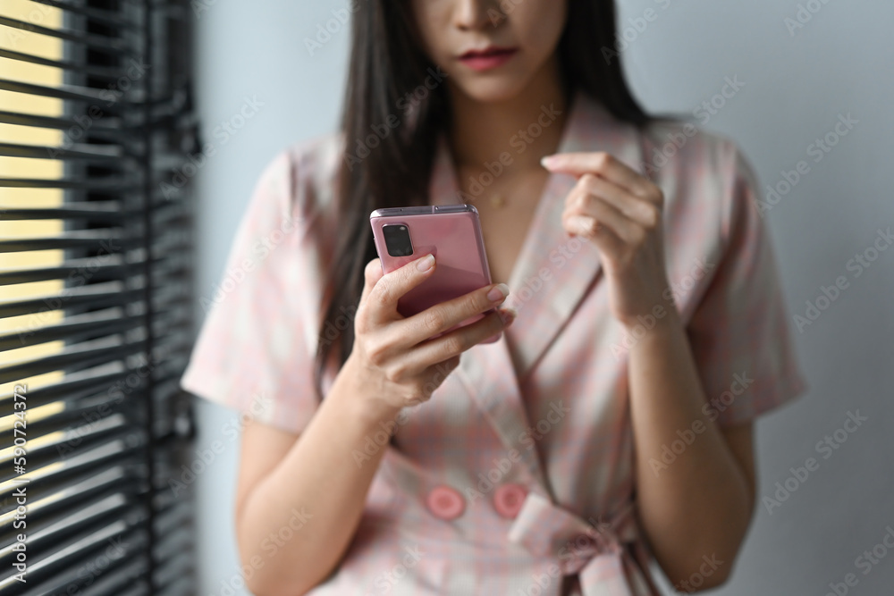 Young woman in stylish dress standing near window and using smart phone.
