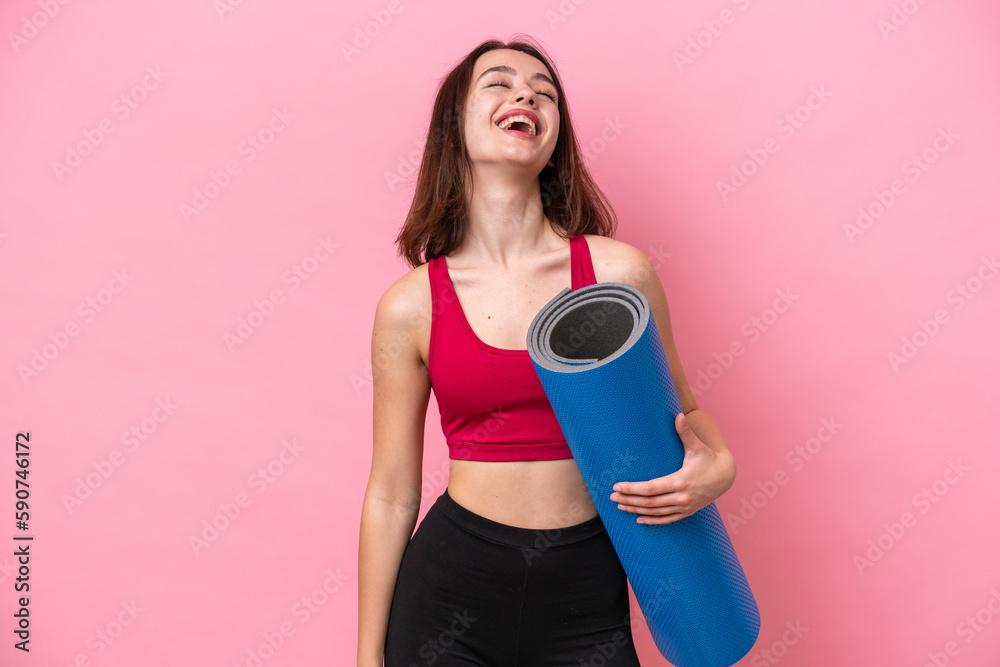 Young sport Ukrainian woman going to yoga classes while holding a mat isolated on pink background laughing