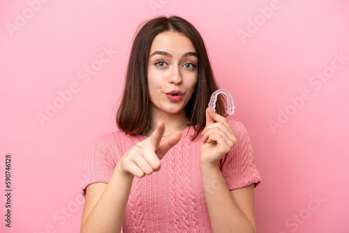 Young Ukrainian woman holding invisible braces isolated on pink background surprised and pointing front