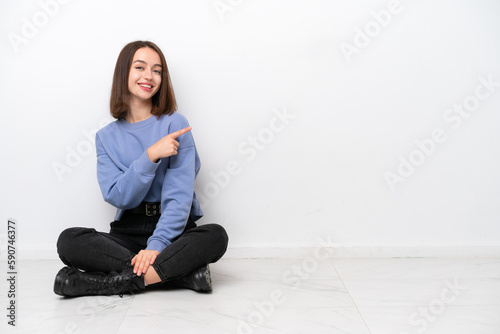 Young Ukrainian woman sitting on the floor isolated on white background pointing to the side to present a product