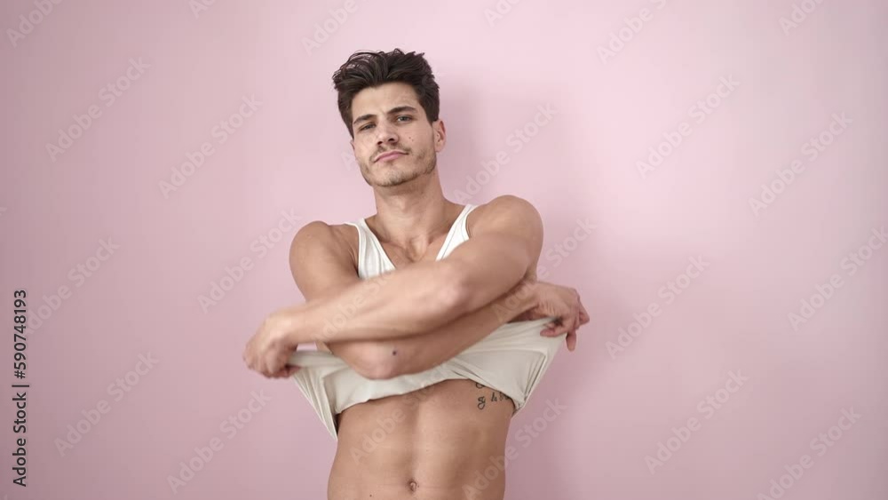 Young hispanic man smiling confident taking off t shirt over isolated pink background