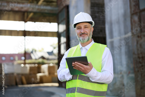 Portrait of the architect in white hard hat and reflective vest standing, working on digital tablet at building site. High skilled mature builder man using modern gadgets during construction work.
