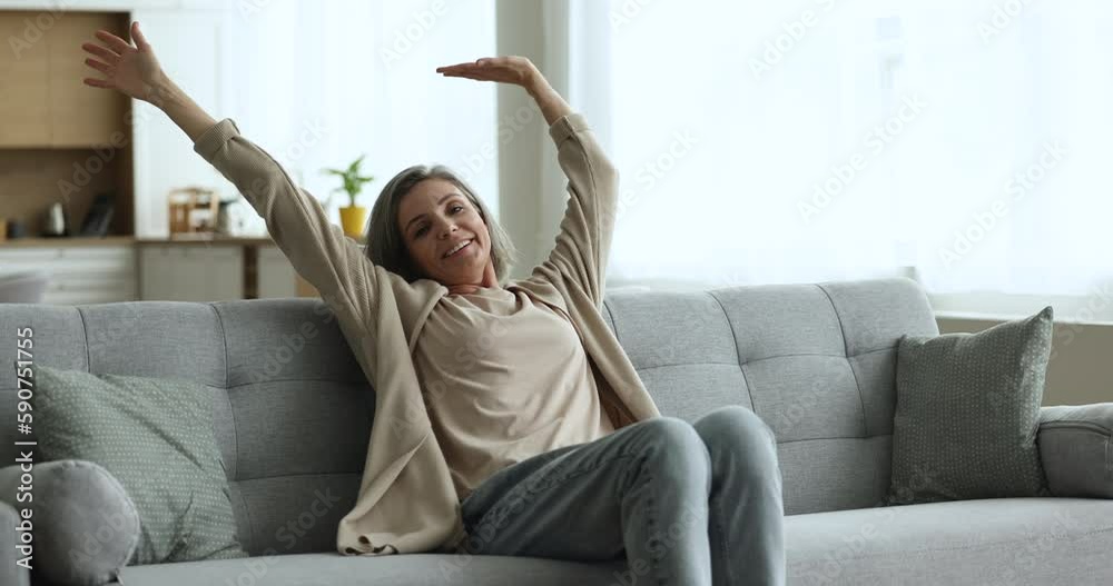 Cheerful middle aged woman walking to couch, sitting down, smiling, relaxing, stretching body with closed eyes, looking away in deep thoughts, thinking, dreaming