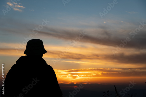 Silhouette of a man wearing a hat standing at a hill and enjoying sunset views. Sun setting and throwing yellow and orange colors into the sky. Sky and clouds look colorful during sunset in india.