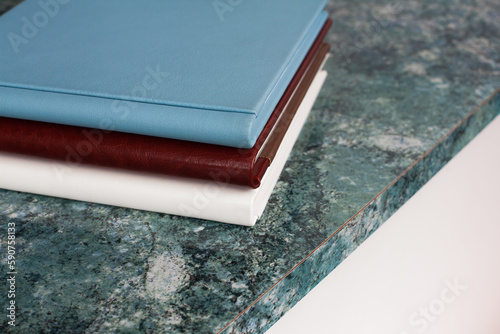 Three stylish photobooks with leather covers, white, burgundy and blue, of different thicknesses, lie on a textured blue surface in a room.