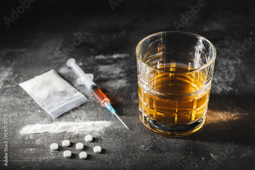 Alcohol drink in a glass, syringe with a dose of drugs, white pills and narcotics powder in a transparent bag on dark background. Concept of addiction, abuse and bad habits