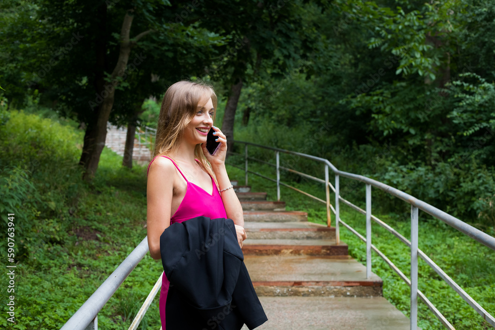 A slender young girl with long blond hair, in a pink dress, stands on the stairs at the railing, in the alley of the park, and talks on the phone, laughs. He holds a black jacket in his hands.