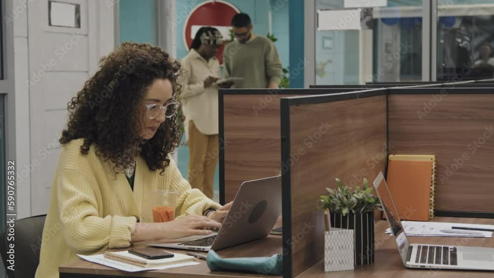 Medium shot of Caucasian woman with curly hair, in glasses and yellow cardigan sitting at desk in cubicle in office, working on laptop and snacking on carrots, and diverse colleagues in background
