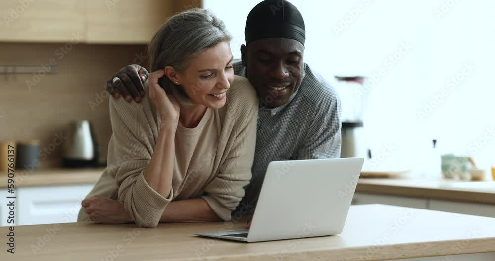 Happy diverse millennial dating couple using laptop in home kitchen, shopping on Internet together, booking hotel for vacation, discussing online purchase, talking, smiling, laughing, hugging