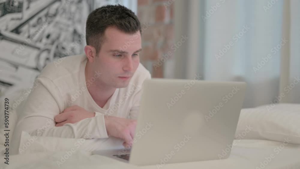 Young Man Working on Laptop while Lying on Stomach in Bed Young Man Working on Laptop while Lying on Stomach in Bed