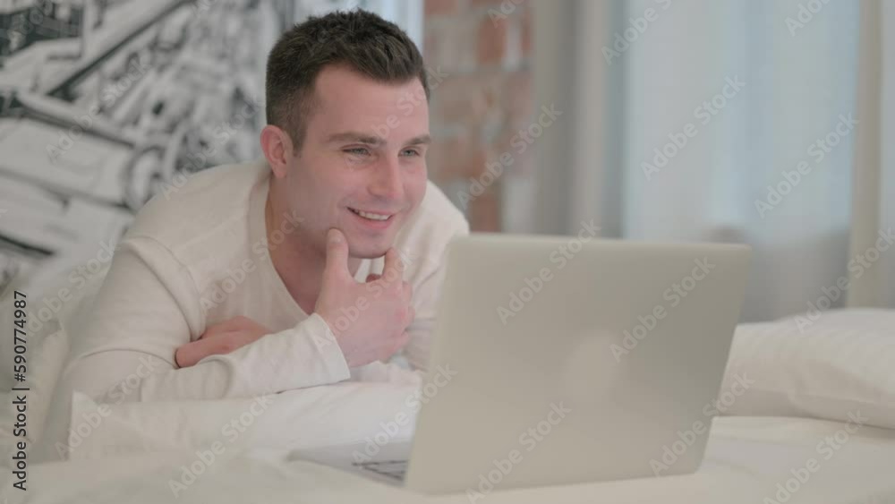 Young Man Doing Video Chat on Laptop while Lying on Stomach in Bed Young Man Doing Video Chat on Laptop while Lying on Stomach in Bed