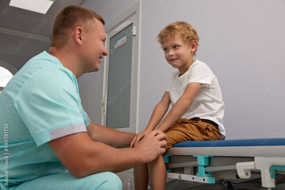 Obraz premium Kind doctor cutely talks with a small patient about the upcoming examination, calms the child. The doctor squatted down next to the couch on which the boy was sitting.