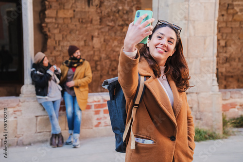 On foreground one young caucasian woman taking a selfie portrait with a mobile phone outdoors, at background a couple talking. isolated tourist lady shooting a photo wearing coat and backpack. High