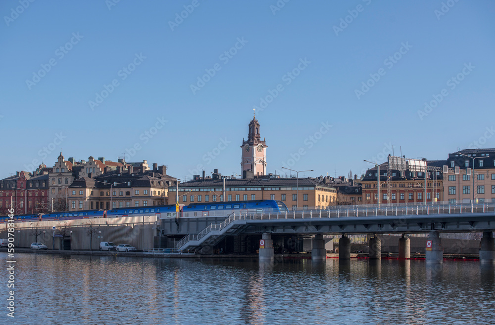 A train bridge at the bay Riddarfjärden, a commuting a blue train, the old town Gamla Stan and the church Tyska kyrkan, a sunny spring morning in Stockholm