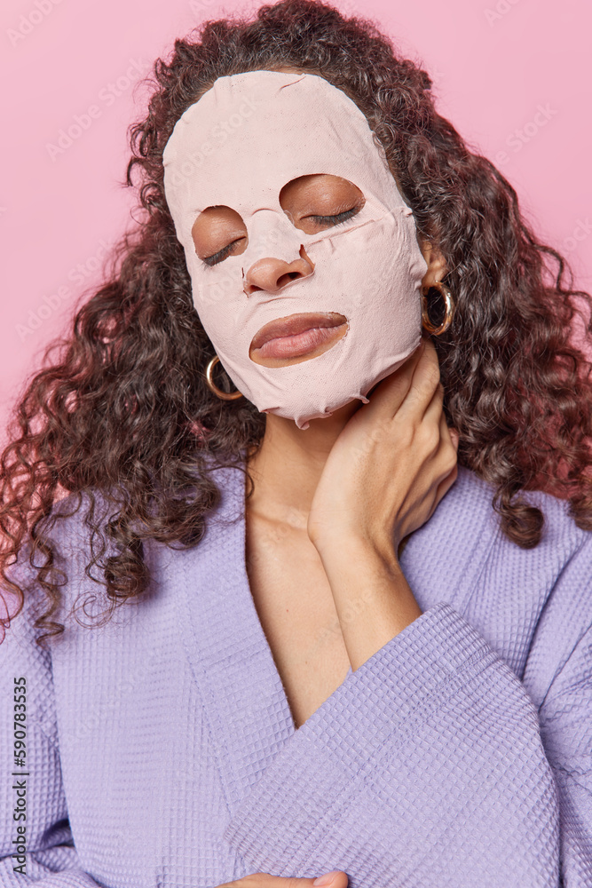 Photo of curly haired woman touches neck keeps eyes closed applies sheet facial mask undergoes beauty procedures dressed in dressing gown isolated over pink background. Pampering procedures. Photo of curly haired woman touches neck keeps eyes closed applies sheet facial mask undergoes beauty procedures dressed in dressing gown isolated over pink background. Pampering procedures.