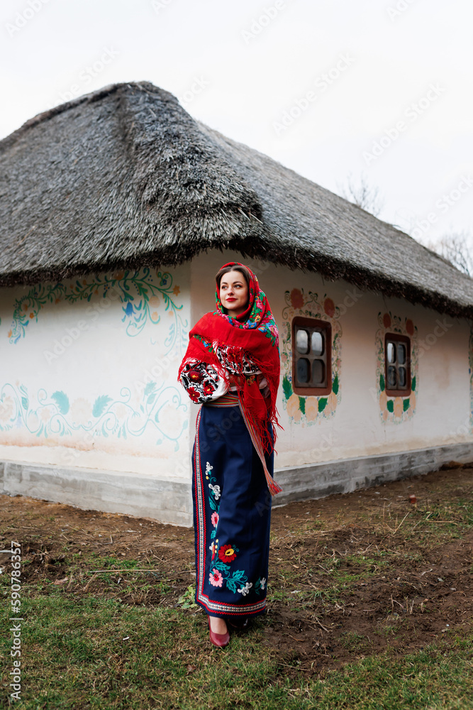 Charming woman in traditional ukrainian handkerchief, necklace and embroidered dress standing at background of decorated hut. Ukraine, style, folk, ethnic culture Charming woman in traditional ukrainian handkerchief, necklace and embroidered dress standing at background of decorated hut. Ukraine, style, folk, ethnic culture