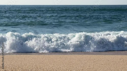 Straight on view of waves breaking on the shore of a sandy beach in spring time in Portugal. Static shot, parallel to the shoreline. Bright sunny day.