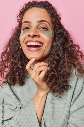 Vertical shot of cheerful woman smiles broadly has white teeth applies patches under eyes to moisturise delicate skin enjoys beauty daily procedures dressed in bathrobe isolated over pink background
