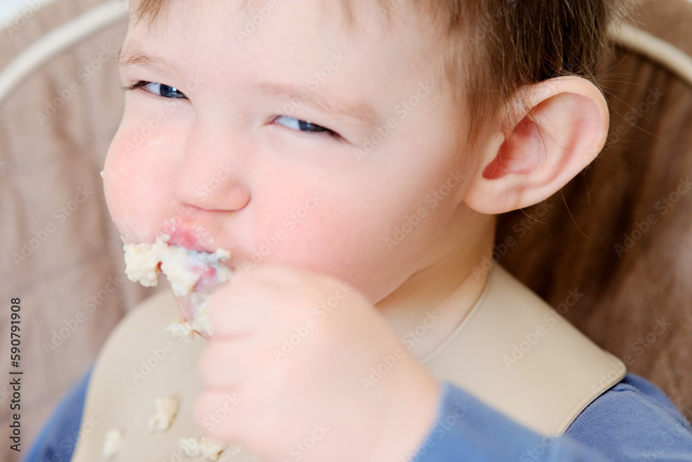 A happy child eats porridge with a spoon while sitting on a high chair. Baby in a bib eats oatmeal porridge himself. Kid aged about two years (age one year nine months) A happy child eats porridge with a spoon while sitting on a high chair. Baby in a bib eats oatmeal porridge himself. Kid aged about two years (age one year nine months)
