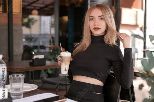 Portrait of young woman with red lips wears black clothes and holds cup of cappuccino. Young attactive girl in cafe. photo