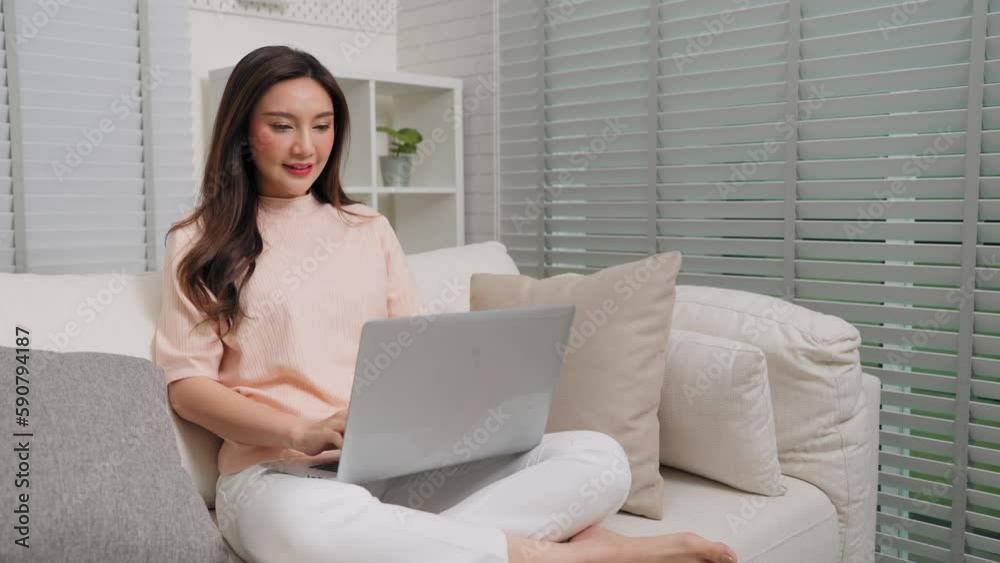 Smiling young Asian woman using laptop and sitting on couch at home, Freelancer female working on computer in living room Smiling young Asian woman using laptop and sitting on couch at home, Freelancer female working on computer in living room